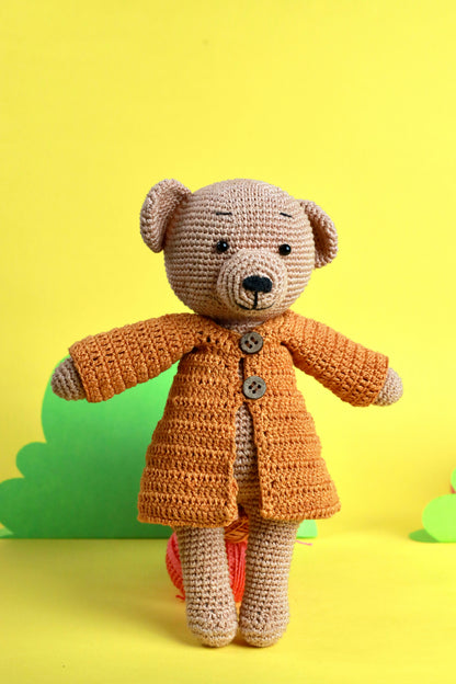 A handcrafted crochet teddy bear with a beige body and an orange coat, standing against a yellow background.