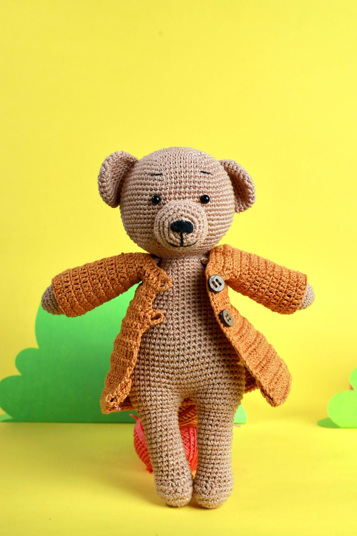 A handcrafted crochet teddy bear with a beige body and an orange coat, standing against a yellow background.